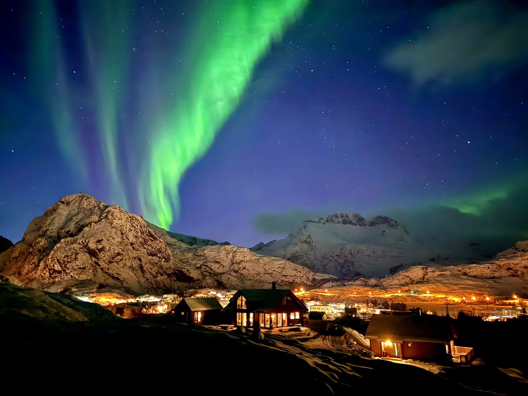 Northern lights over the cabin cluster on a winter night, with snow-covered mountains behind